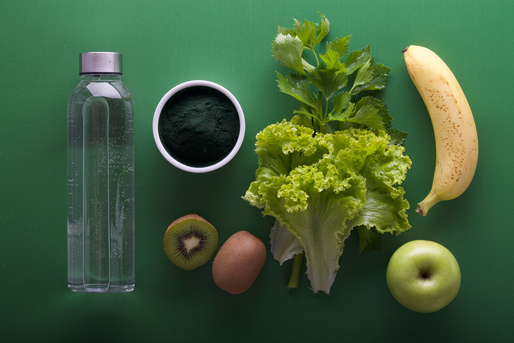 Picture of a water bottle, spirulina, one and a half kiwi, lettuce, and apple and a banana on a green background.