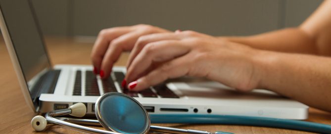 Picture of hands typing on a laptop with a stethoscope on the table.