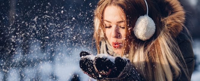 Image of a woman in a coat and earmuffs blowing snow from her mittened hands.